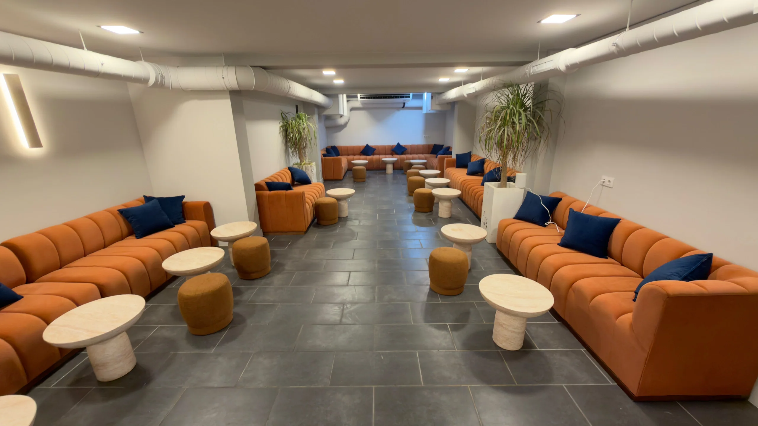 Sala principal de Cloud 420 Weed Club, club cannábico en Port d'Andratx (Mallorca): bancos corridos naranjas con cojines azules, mesas redondas de travertino y suelo de piedra gris
